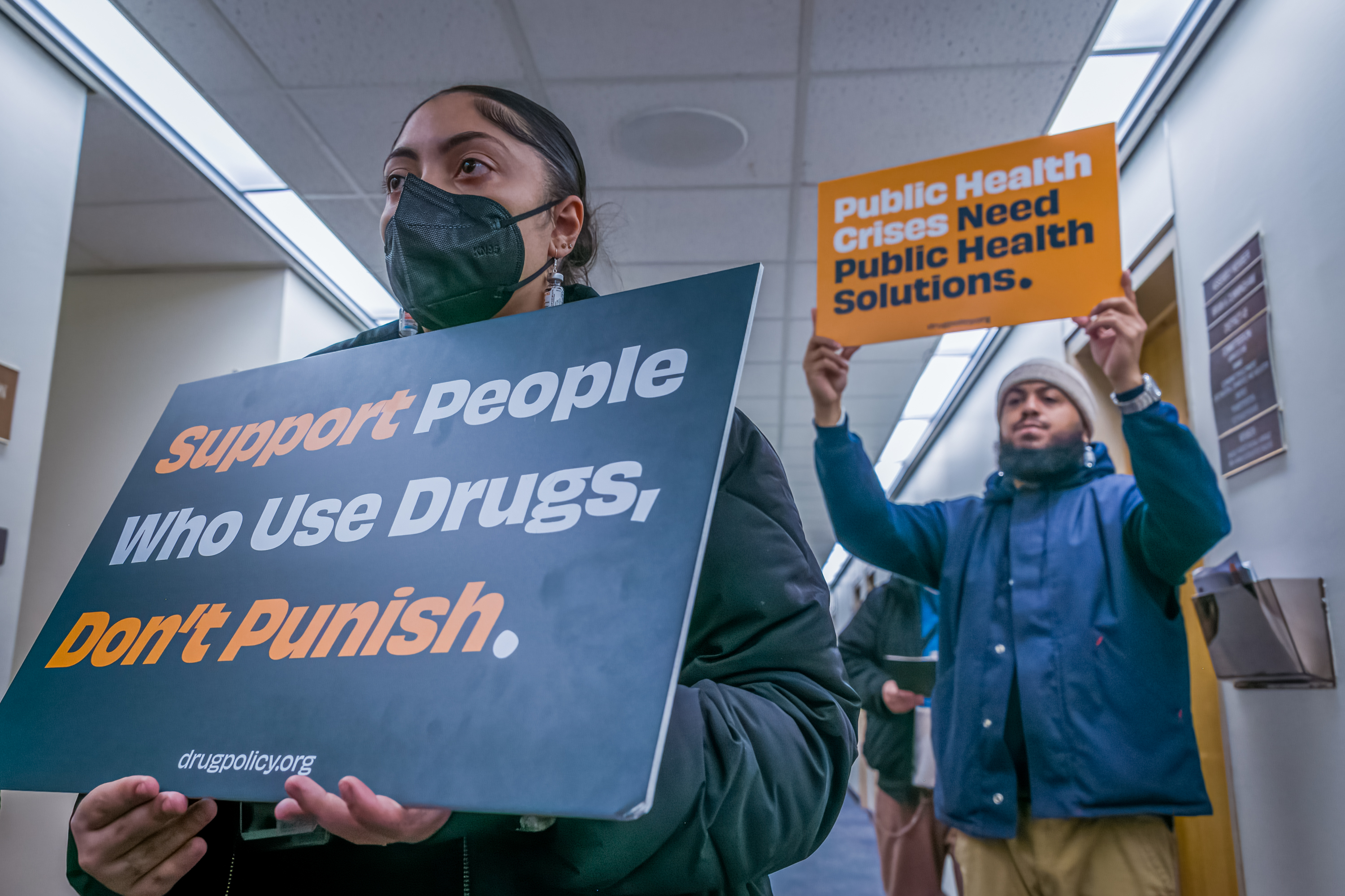 Two people hold orange and green signs. The visible sign reads "Support people who use drugs, don't punish"